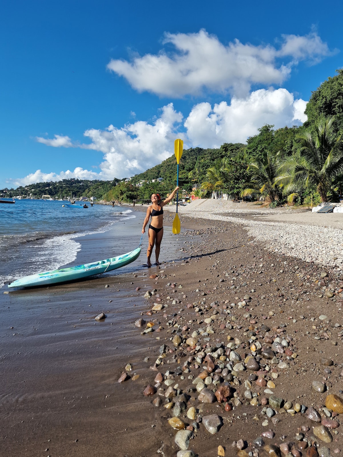 A person stands on a sandy beach, holding a yellow paddle above their head. A kayak rests on the shore beside them, and a backdrop of lush green hills and scattered clouds fills the scene. The water is calm and reflects the clear blue sky.
