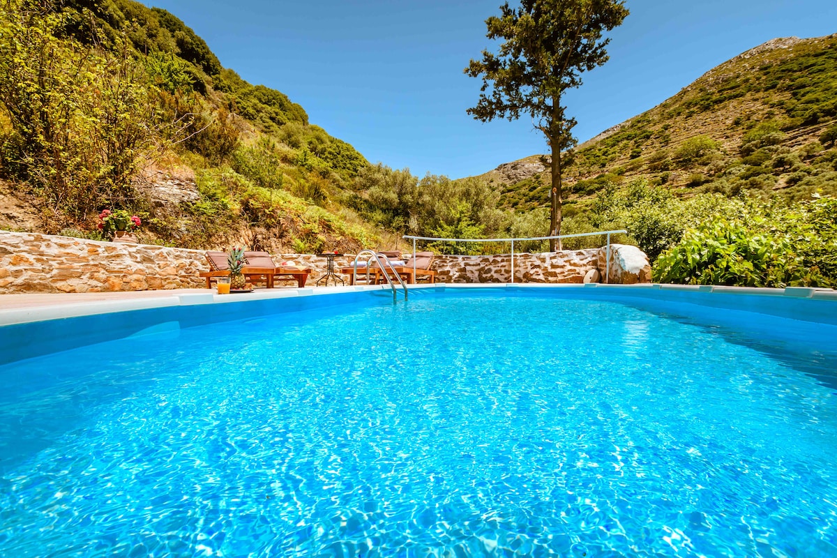 A tranquil swimming pool reflects the clear blue sky, surrounded by lush greenery and distant hills. Sun loungers are positioned nearby for relaxation, creating a serene outdoor space. Natural stone walls enhance the landscape, contributing to the peaceful ambiance.