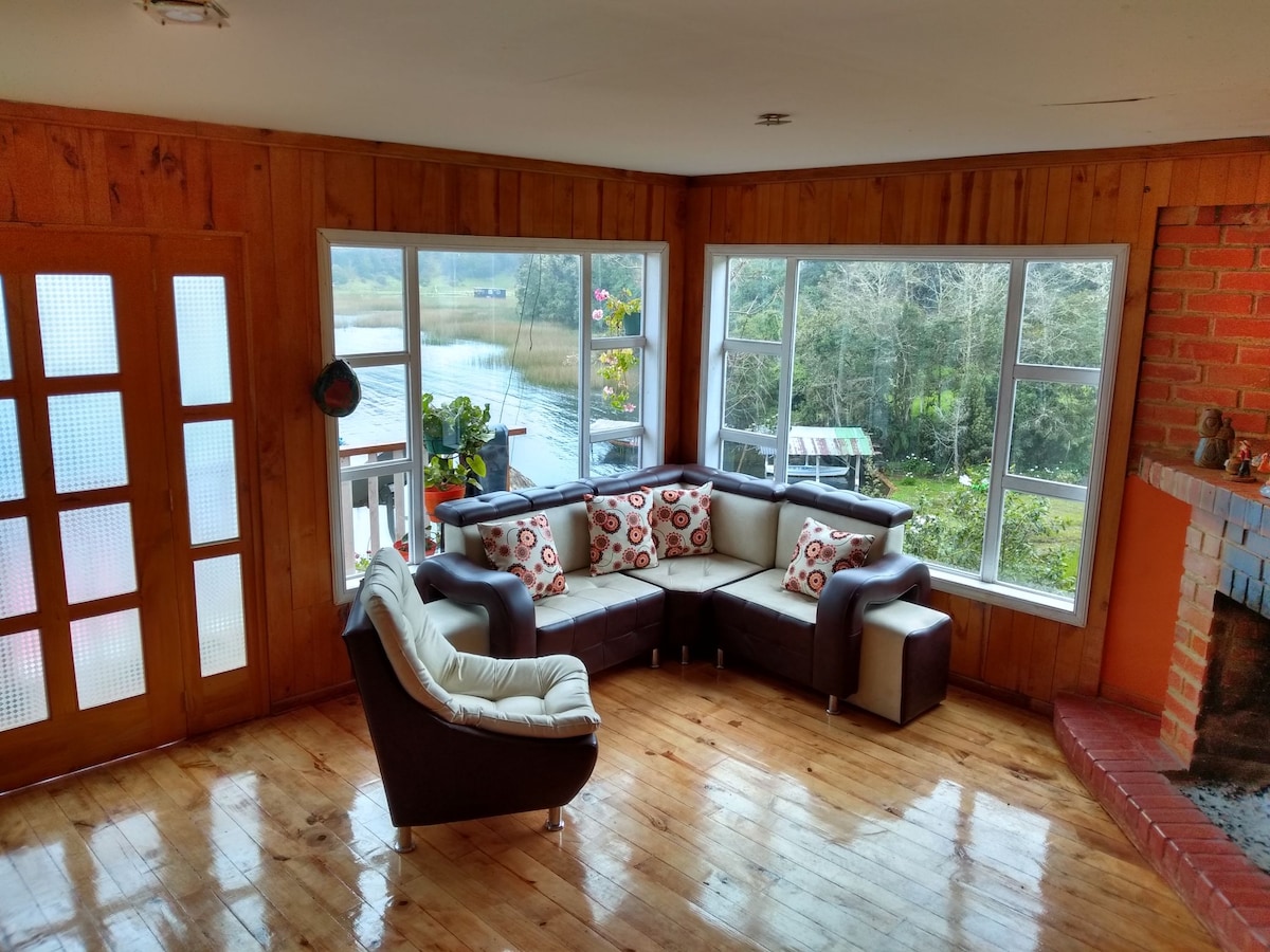 A living area features a curved sectional sofa adorned with patterned cushions, positioned near large windows offering views of the surrounding landscape. Natural light fills the space, highlighting the wooden walls and polished floor. A rustic brick fireplace adds warmth to the ambiance.