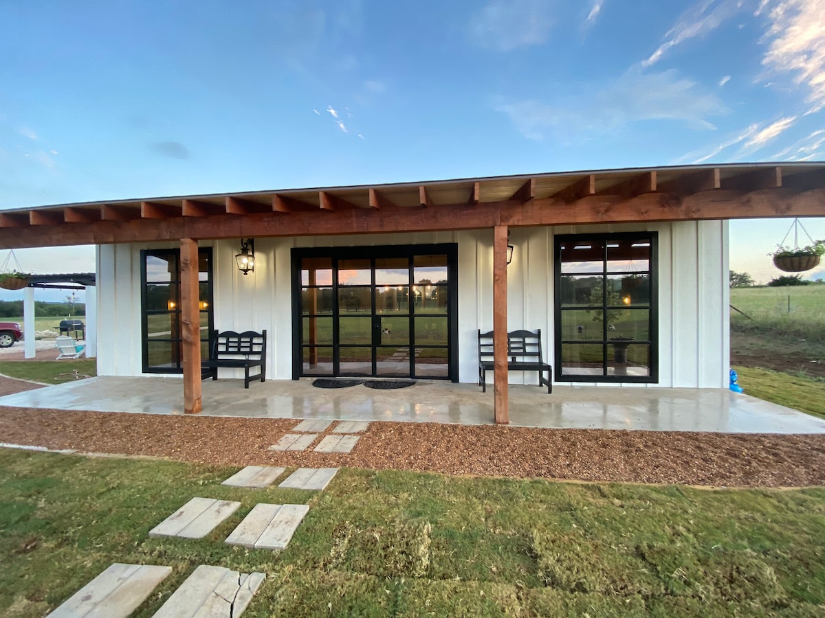 The inviting exterior features a wide porch with two benches and spacious glass doors, allowing for natural light to flow inside. The surrounding lawn is well-maintained, accented by stone pathways and decorative elements, with open fields visible in the background.