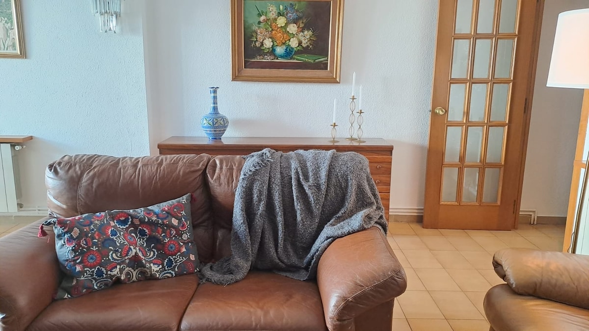 A comfortable leather sofa is seen, draped with a soft gray throw. An artwork featuring floral arrangements adorns the wall behind the sofa. A wooden cabinet and a door with multiple glass panels are present, adding to the inviting living space.