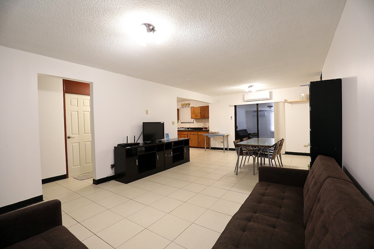 A spacious living area is presented, featuring a dark brown sofa and a sleek black television stand. The open kitchen is visible in the background, showcasing wooden cabinetry and counter space. A modern dining table with several chairs occupies the central area, complemented by tile flooring.
