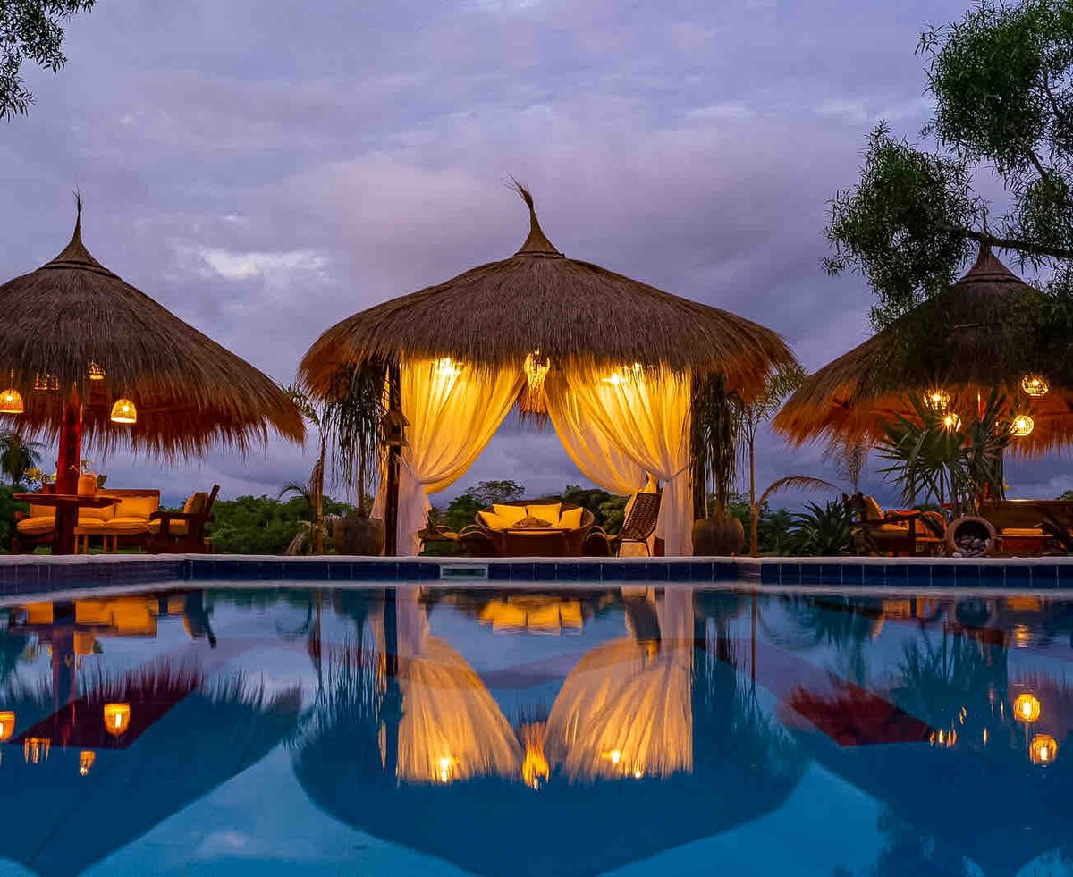 A serene outdoor area features three thatched-roof cabanas with soft, flowing curtains. The shimmering pool reflects the calming scene, illuminated by warm lights. Surrounded by lush greenery, the inviting space suggests relaxation and comfort in nature.