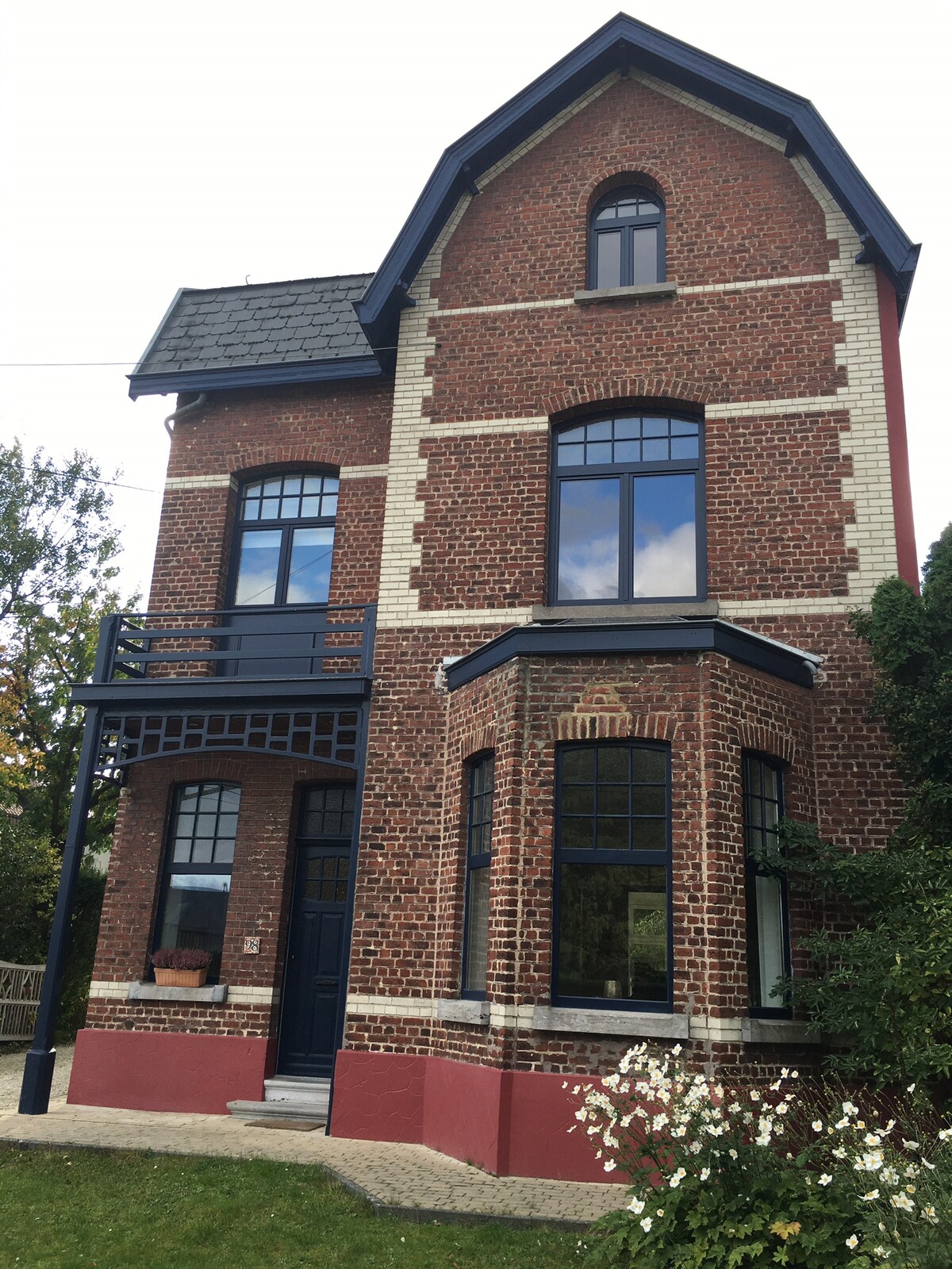 A three-story house with a brick façade is presented, featuring large windows framed in black. A covered porch provides an entrance, and white flowers are visible in the foreground, enhancing the greenery around the home. The architectural details reflect a charming design.