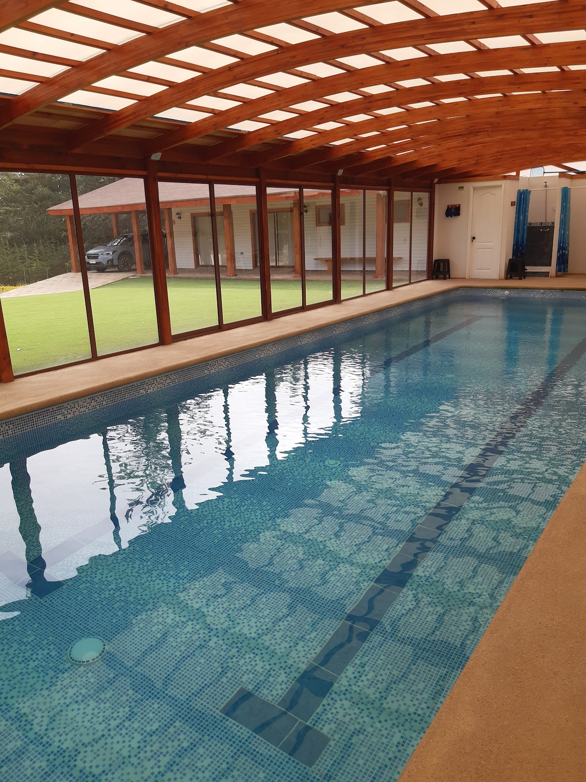 An indoor heated pool is framed by large glass panels, allowing ample natural light to illuminate the clear water. Wooden beams form an overhead structure, complementing the tranquil environment. In the background, a grassy area and the exterior of the nearby house are visible.