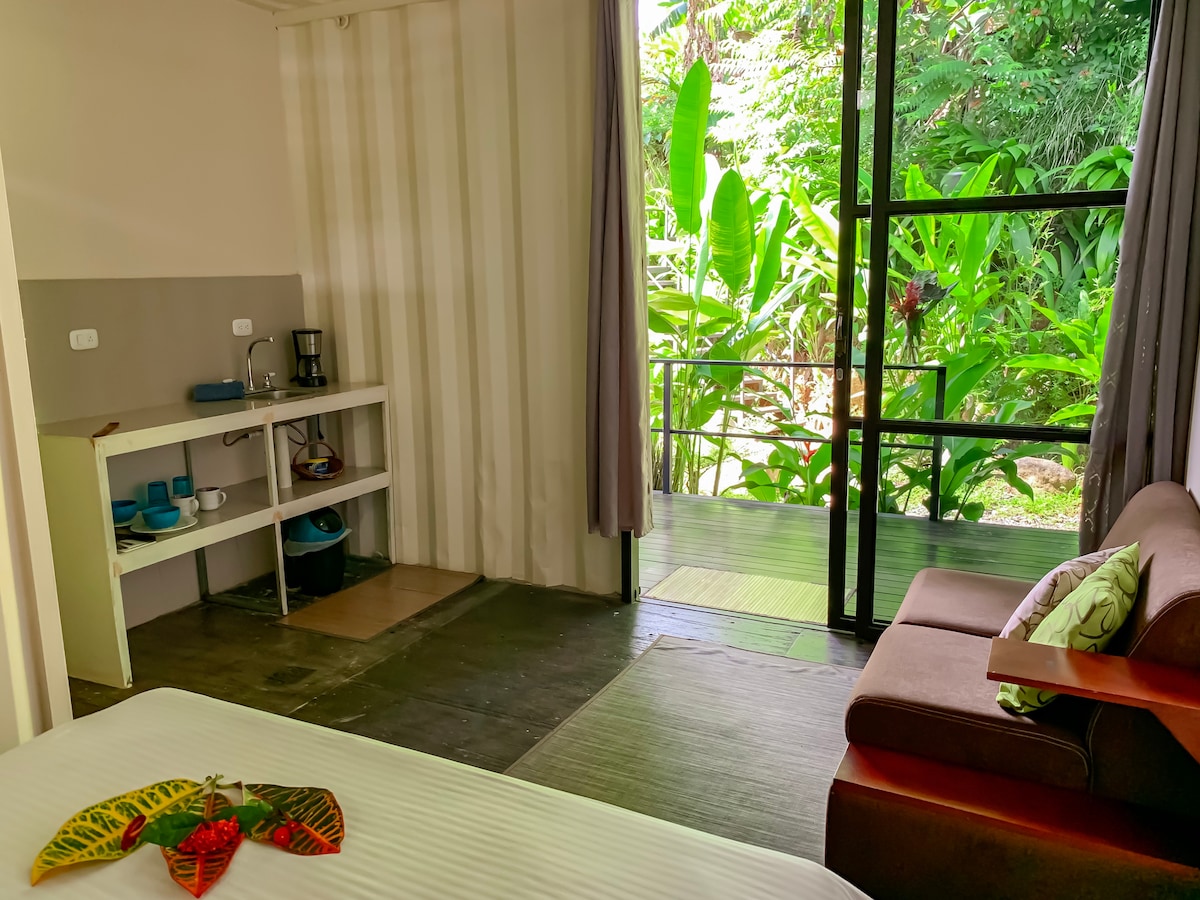 A bright room features a spacious bed with decorative leaves placed on top. A small kitchenette is visible with essential utensils and a coffee maker. Large glass doors lead to a private outdoor space filled with lush greenery.