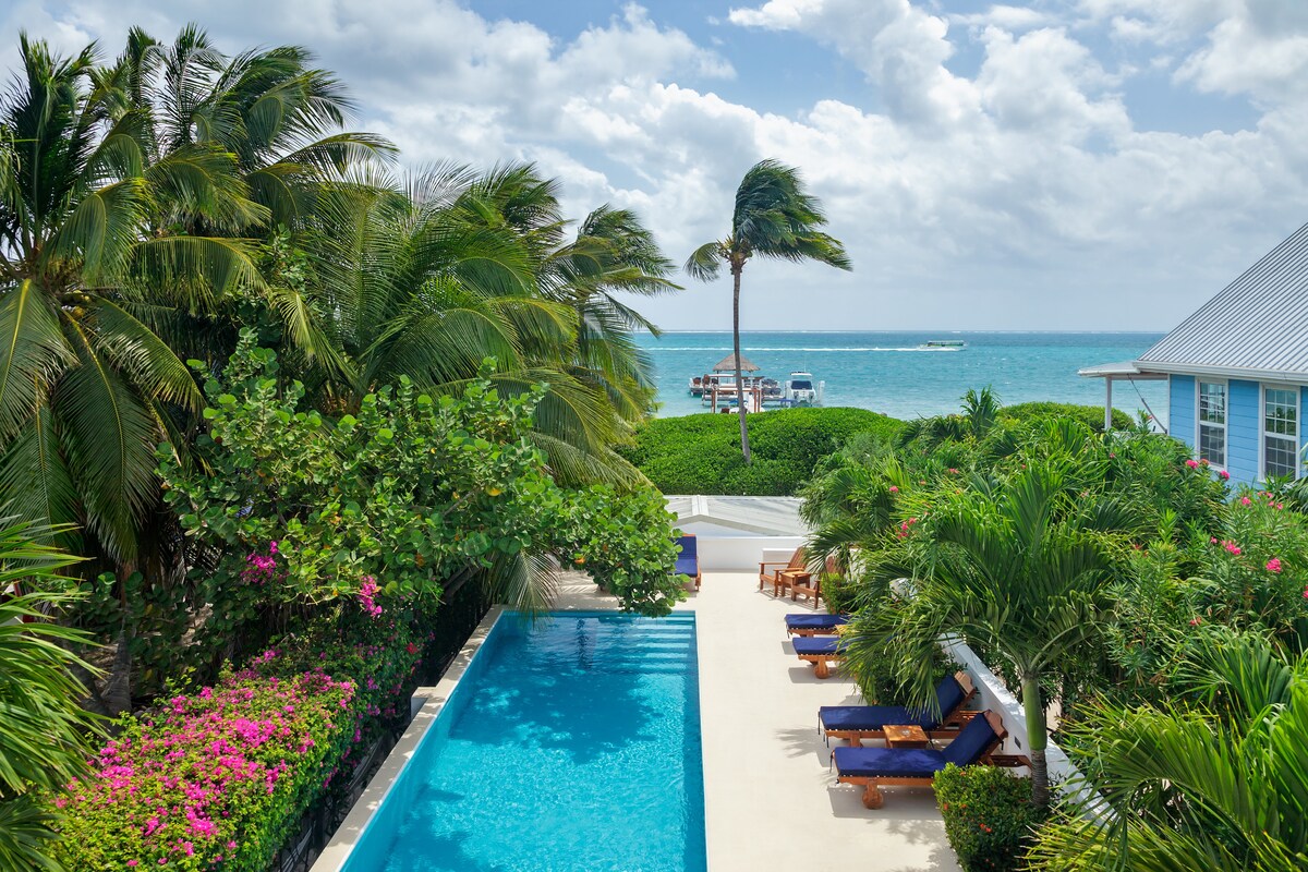 A serene outdoor area features a bright blue swimming pool bordered by lush greenery and colorful flowering plants. Sun loungers are arranged poolside, with a view of the ocean visible in the distance under a partly cloudy sky.
