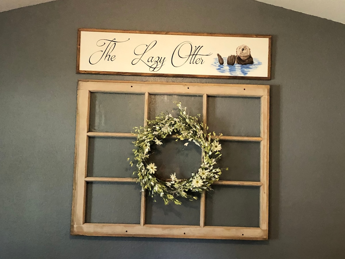 A charming wall decor features a rustic wooden frame resembling a window, adorned with a round floral wreath. Above it, a sign displaying the name 'The Lazy Otter' is showcased, enhancing the welcoming ambiance of the space.
