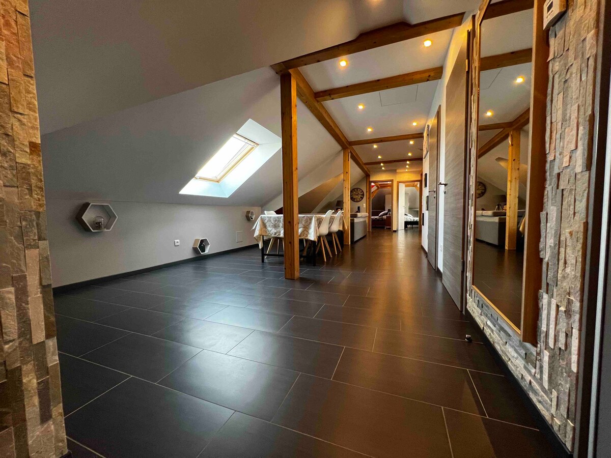 A spacious open area is highlighted by sleek dark tiled flooring and wood-beamed ceilings. Natural light enters through a skylight, illuminating the dining table with chairs arranged neatly. Visible walls feature a mix of smooth and textured finishes, enhancing the modern aesthetic.