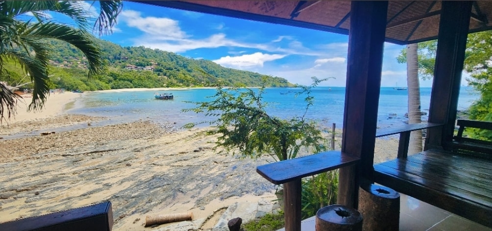 A serene beachfront view is presented, showcasing clear turquoise waters and a sandy shore. Lush green hills rise in the background, while palm trees frame the scene. A wooden structure offers shaded seating, providing an ideal perspective of the tranquil landscape.