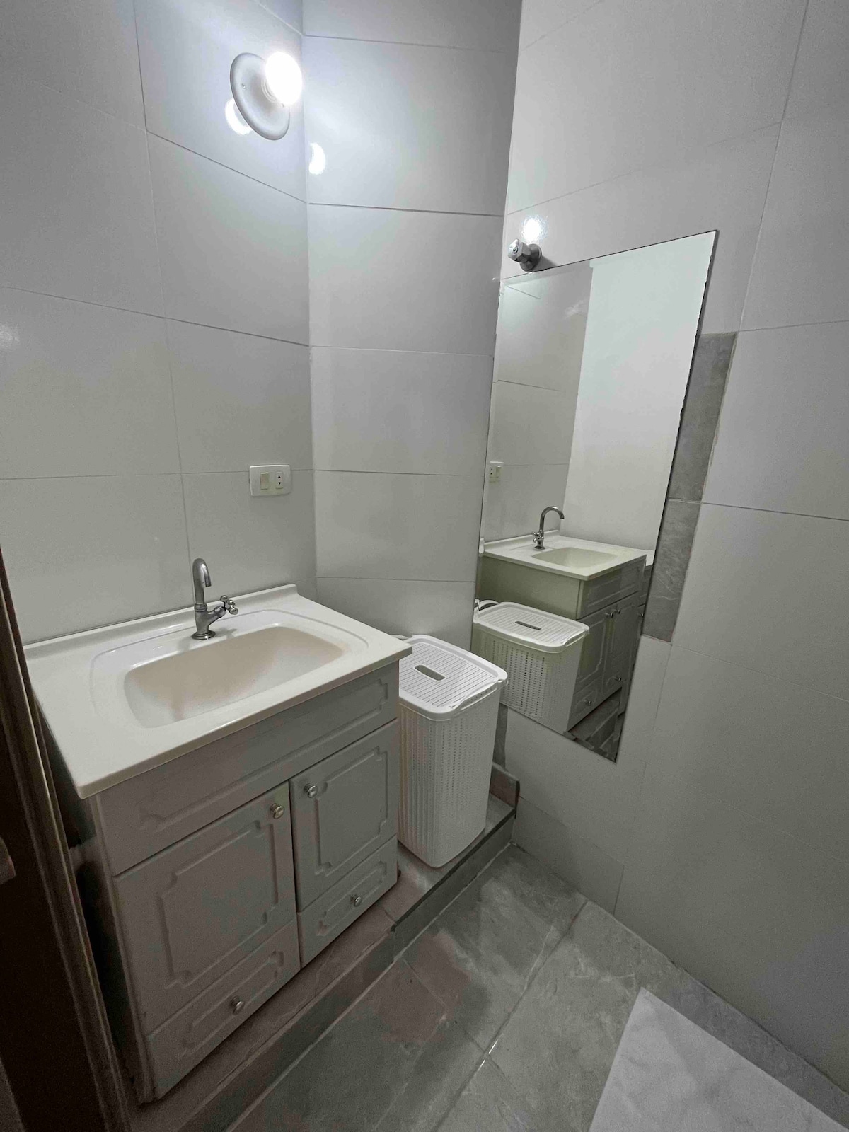 The bathroom features a compact sink area with storage beneath, complemented by a large mirror that enhances the sense of space. A white trash bin is positioned nearby. Soft lighting offers a bright and functional atmosphere.