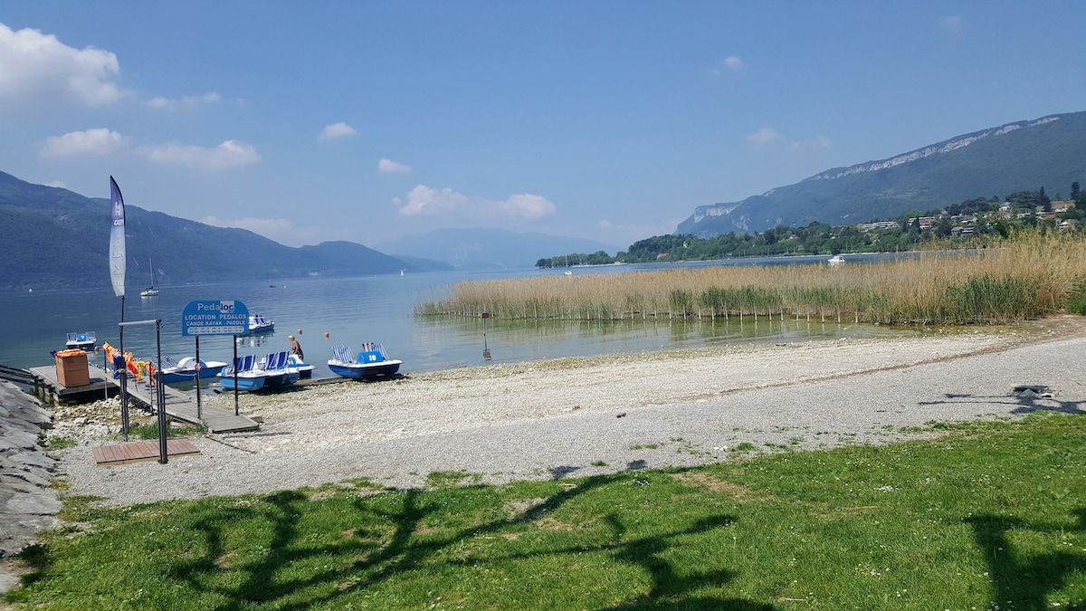 A scenic lakeside view features calm waters bordered by lush greenery. A stony shore is visible, with small boats moored nearby. In the distance, hills rise gently against a partly cloudy sky, creating a peaceful recreational atmosphere.