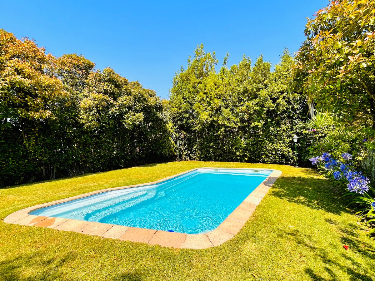 A private swimming pool is nestled within a landscaped garden, surrounded by lush greenery. The clear blue water sparkles under the sunlight, and the pool is framed by a meticulously maintained lawn, creating an inviting outdoor space for relaxation.