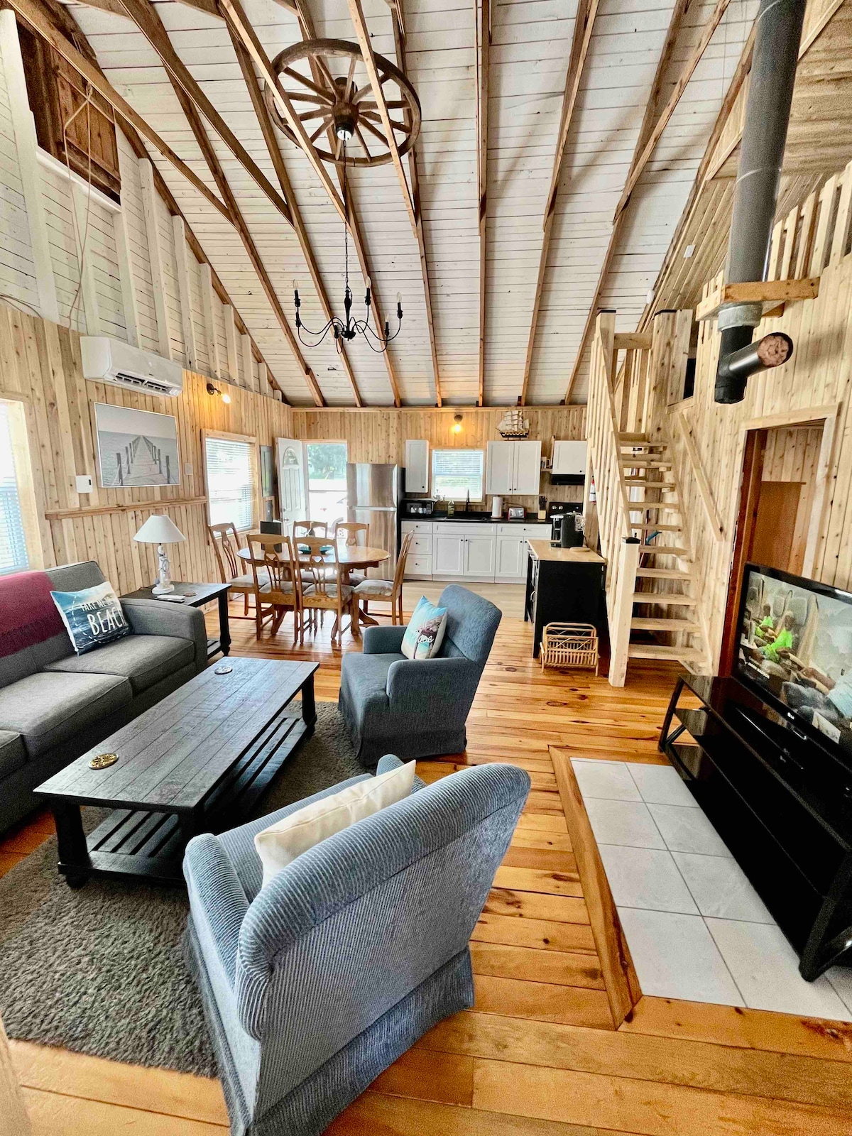 An open-concept living area features a cozy seating arrangement with a dark coffee table and soft furnishings. The exposed wooden beams add character to the space, while a dining table is positioned nearby. A staircase leads to an upper loft area, enhancing the room's spacious feel.