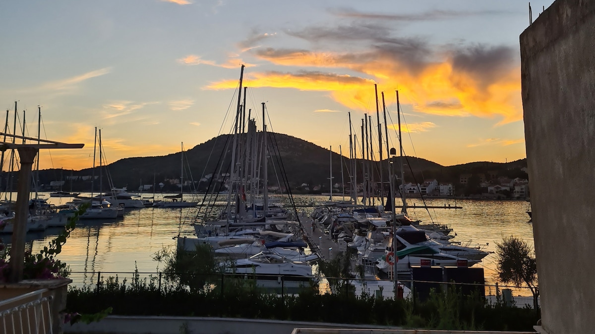 A marina scene is captured at sunset, featuring numerous sailboats lined along a crescent-shaped dock. The water reflects the warm colors of the sky, while hills form a backdrop. Soft clouds add texture to the skyline, enhancing the tranquil ambiance of the setting.