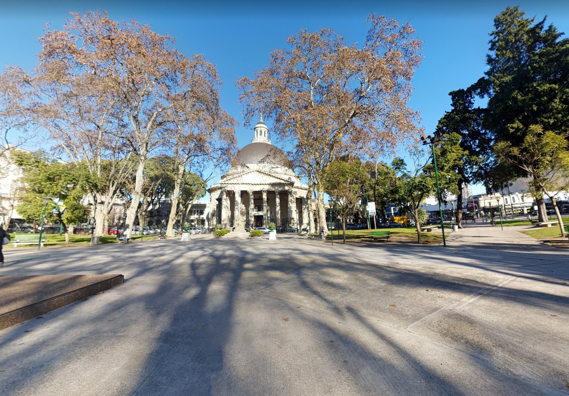 A spacious public park is featured, with a large domed pavilion at the center surrounded by columns. Leafless trees cast shadows on the well-maintained ground, while green foliage is visible in the background. Clear blue skies provide a serene atmosphere.
