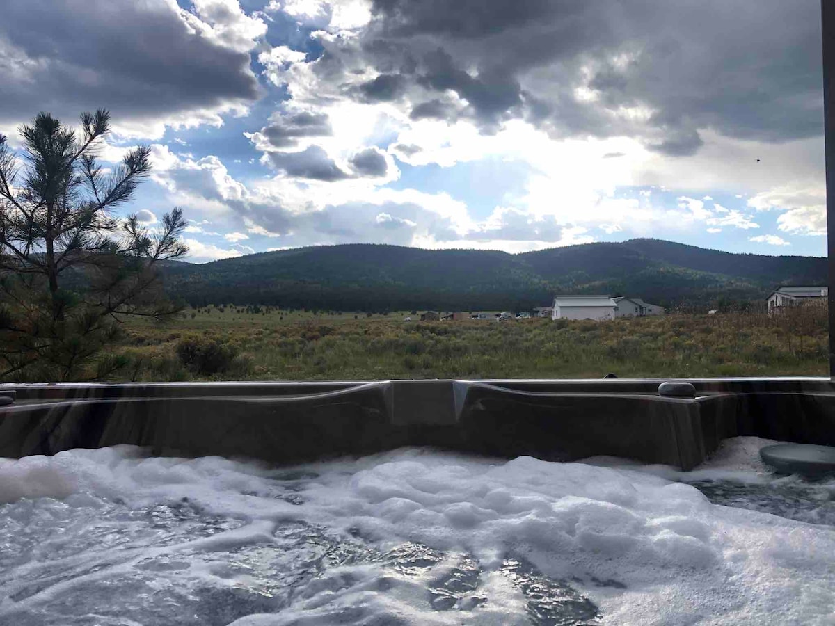 The image shows a hot tub surrounded by nature, with jets creating bubbles in the water. A vast landscape of rolling hills and distant mountains is visible under a partly cloudy sky, adding to the serene environment.