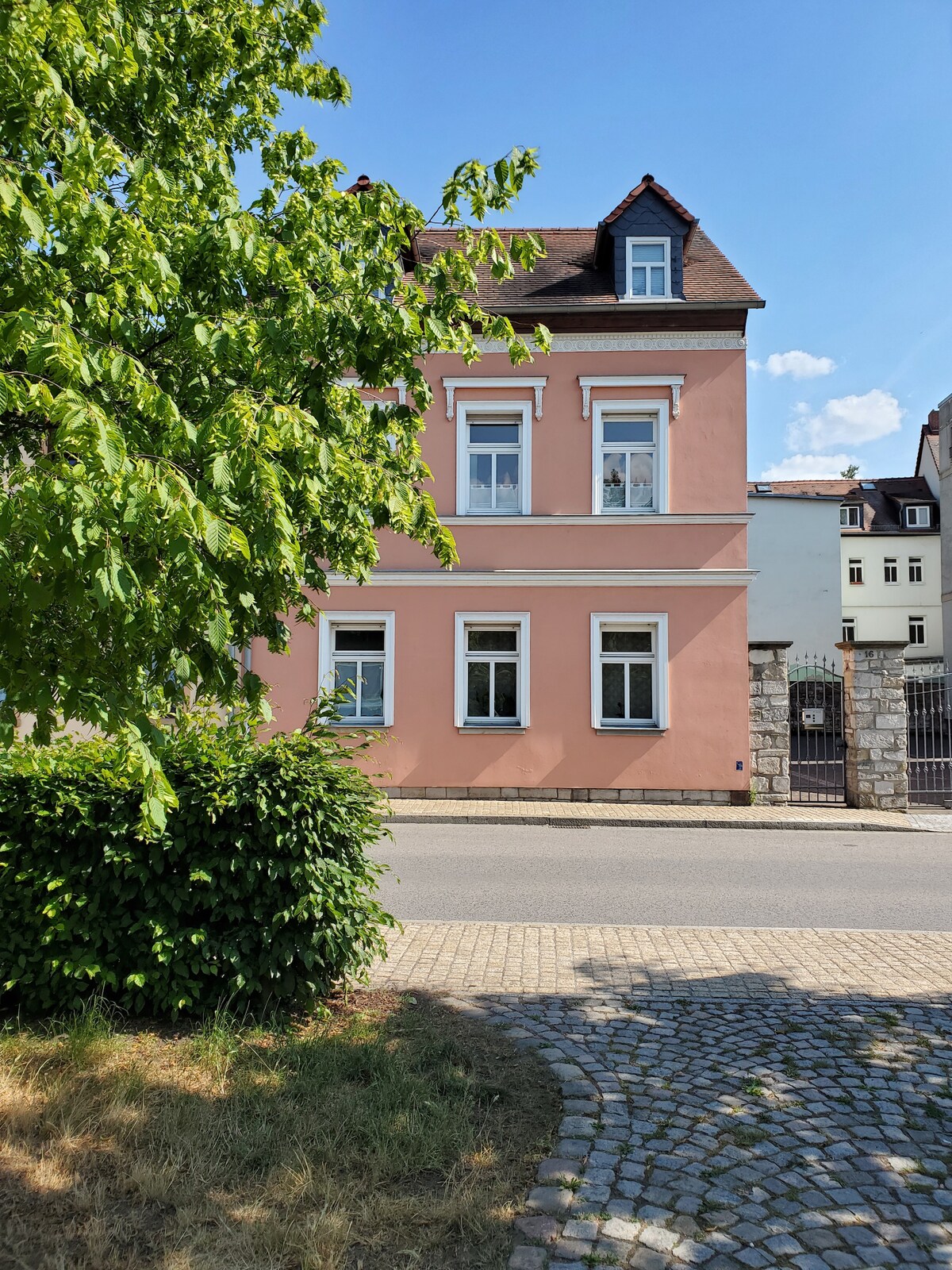 A charming pink building is framed by greenery, featuring multiple windows and a steep roof. The well-maintained exterior is visible from the street, with cobblestones lining the area and nearby structures partially seen in the background.