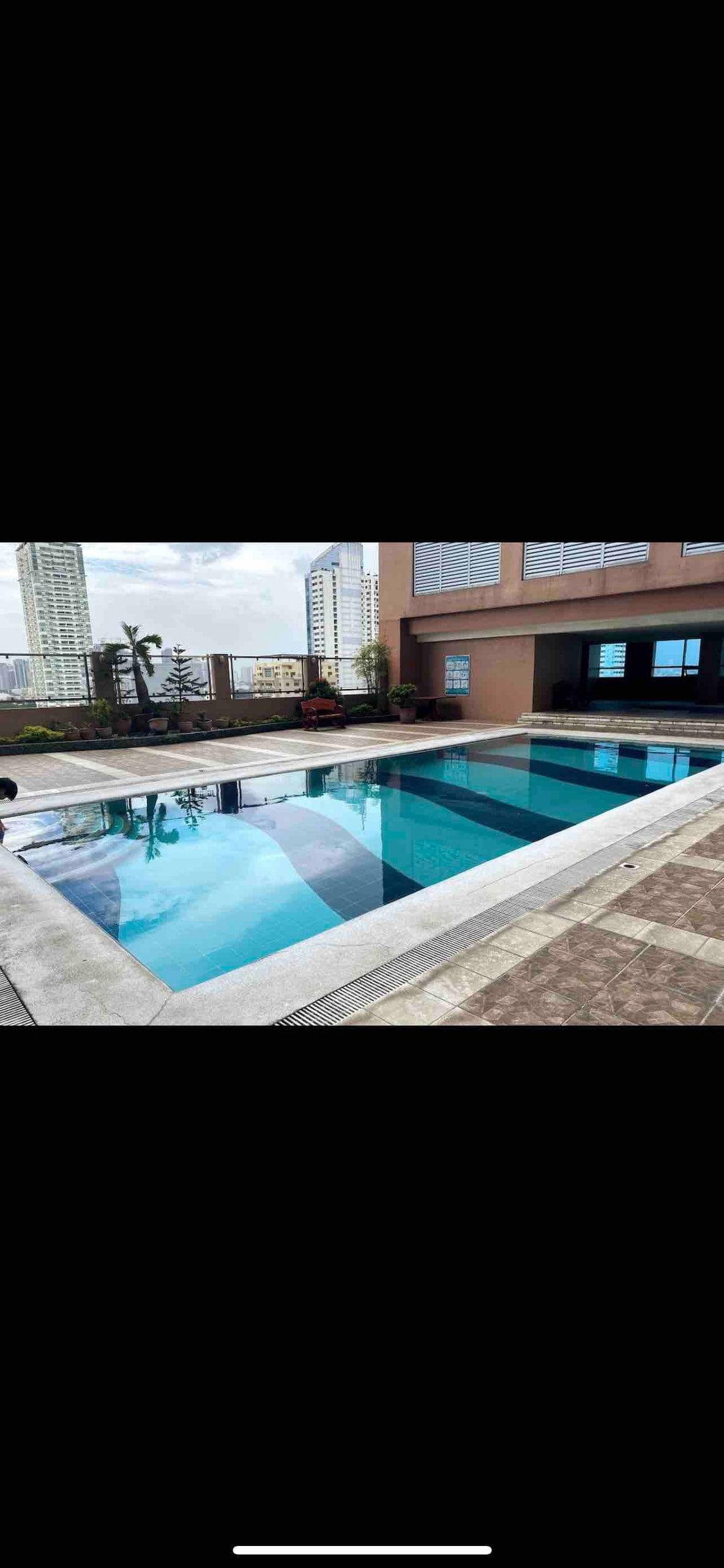 An outdoor swimming pool is presented, bordered by a smooth stone deck. Surrounding structures and palm trees offer a glimpse of urban life, while the calm water reflects the buildings and sky above. Lounge chairs are positioned nearby for guest relaxation.