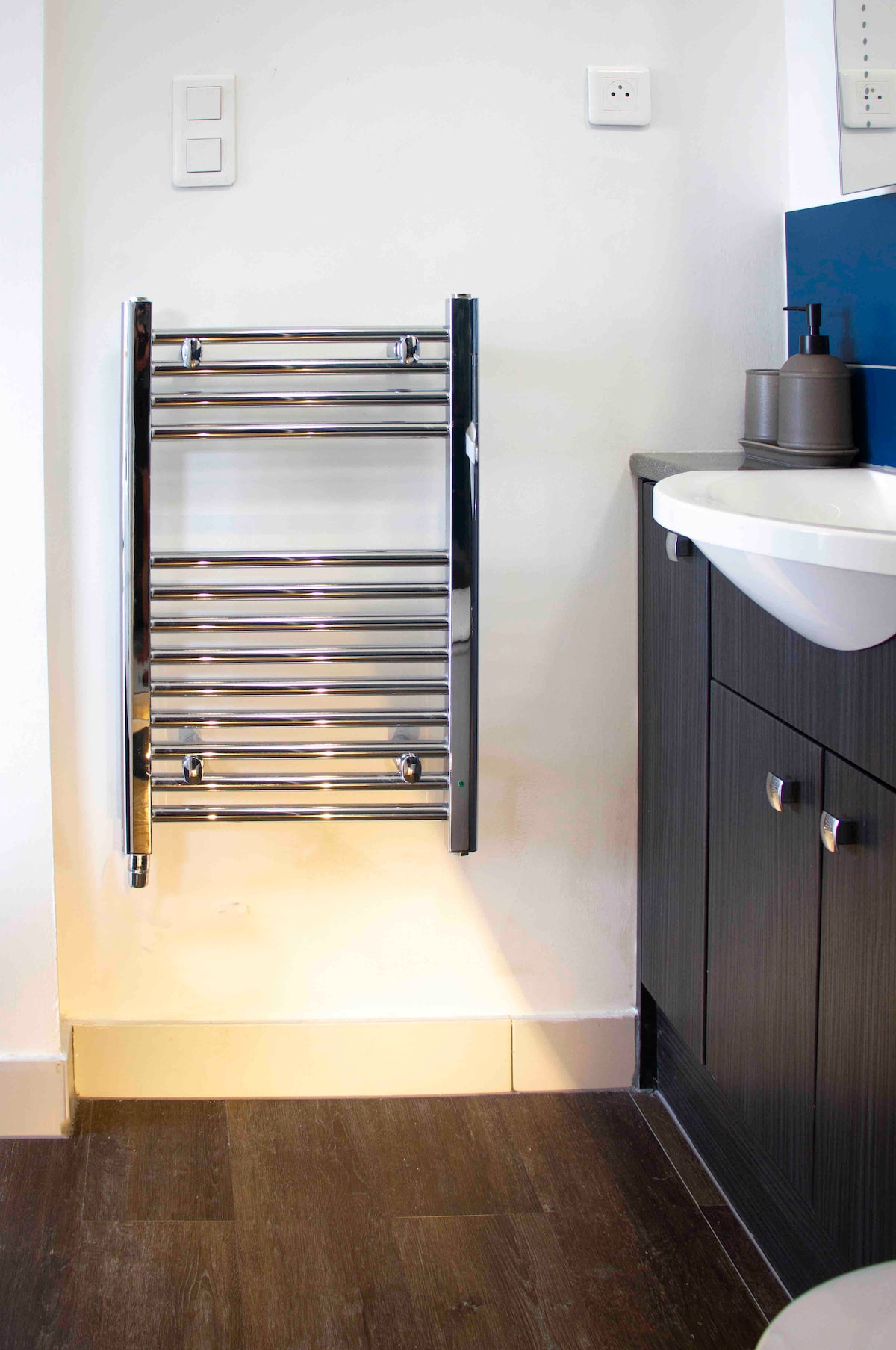A modern towel radiator is mounted on the wall, reflecting light to create a bright, functional space. A sleek vanity cabinet is positioned nearby, featuring dark wood finishes and a simple sink. The floor is laid with dark tiles, complementing the overall design.