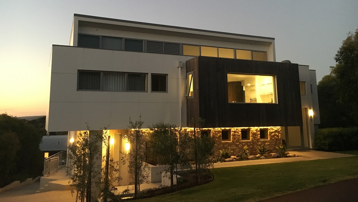 A modern home exhibits a sleek design with a combination of white walls and wooden accents. Outdoor lighting highlights the entryway and landscaped gardens, offering a welcoming appearance during dusk. Large windows provide glimpses of the interior spaces, blending functionality with contemporary style.