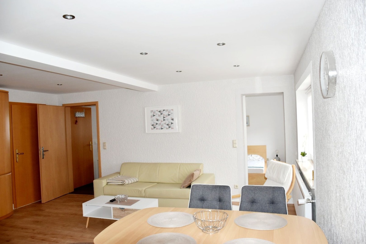 The living area displays a light-colored sofa facing a modern coffee table. A wooden dining table with grey chairs is visible in the foreground. Two wooden doors lead to separate rooms, while a simple wall clock complements the minimalist decor.