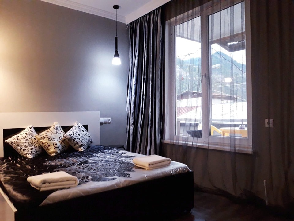 A well-lit bedroom features a spacious bed dressed in dark bedding with decorative pillows. Large windows adorned with sheer curtains allow natural light to filter in, highlighting the soft grey walls. The outdoor view reveals a mountainous backdrop.