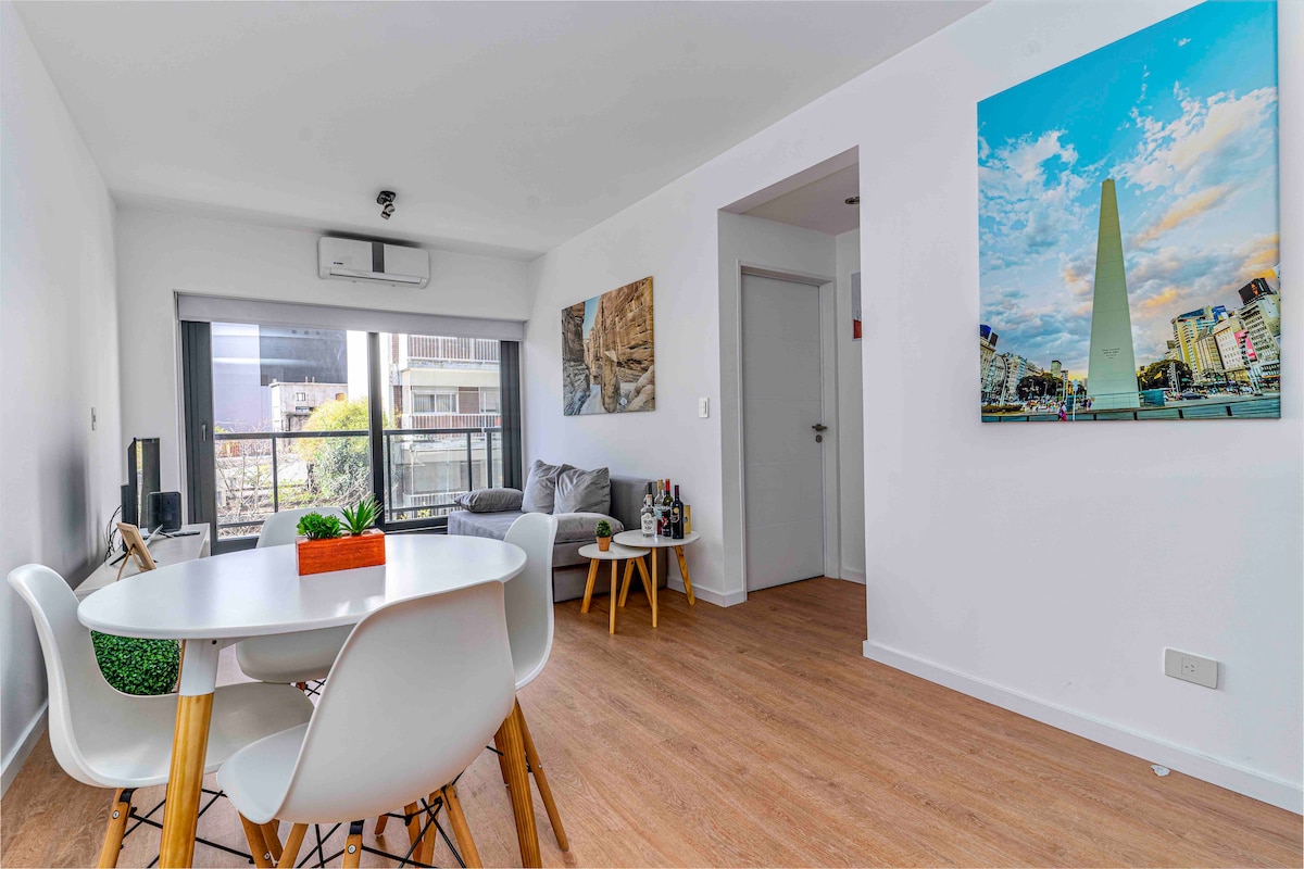 A bright living area features a circular dining table with white chairs. A modern gray sofa is positioned near large windows that allow natural light, with a colorful wall art piece of a cityscape adding a vibrant touch.