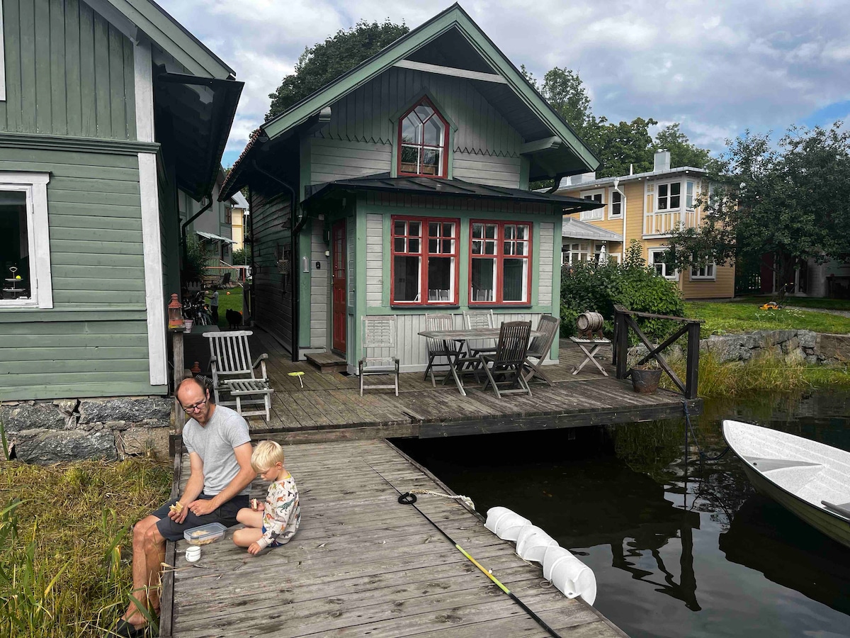 A charming green fishing cabin, dating back to 1882, is featured next to a wooden dock. Outdoor seating is arranged on the deck, overlooking the water. A small child and an adult are seated on the dock, with a rowboat visible nearby.