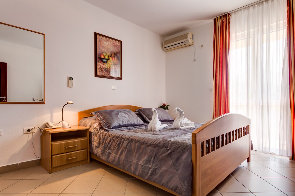 A comfortable studio apartment is displayed with a spacious bed featuring subtle bedding patterns. A bedside table with a lamp is present, along with a decorative painting on the wall. Natural light enters through large windows, complemented by flowing curtains.