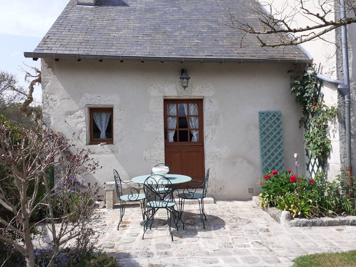 An outdoor dining area features a round table surrounded by wrought-iron chairs, situated on a stone patio. The entrance to a cottage is framed by a wooden door with a window above, complemented by flowering plants and greenery along the walls.