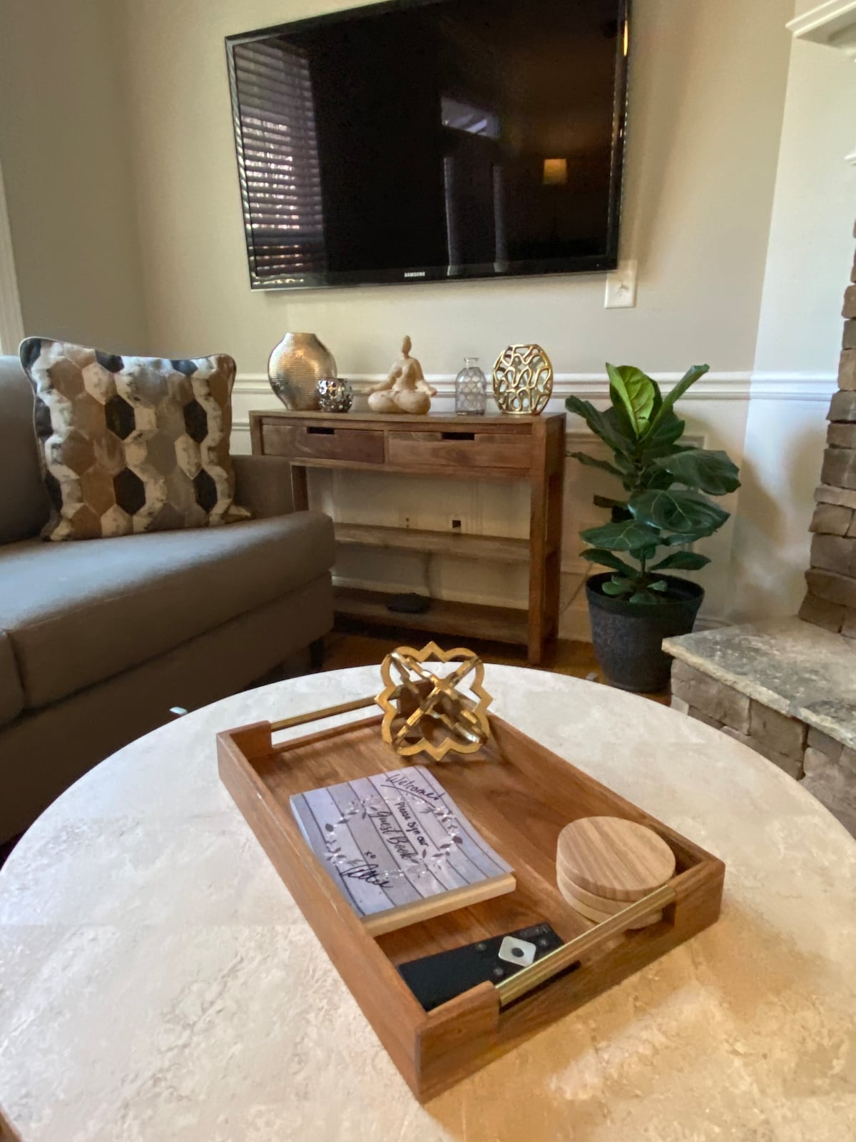 A cozy living area features a light-colored coffee table with a wooden tray holding decorative items and a notepad. A plush gray couch with patterned cushions is positioned next to a wooden console table, while a small plant adds a touch of greenery. A flat-screen television is mounted on the wall.