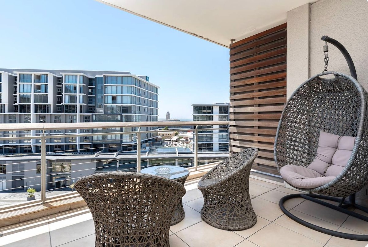 An outdoor balcony area features a hanging chair and two textured, round chairs, surrounded by a railing. The city views showcase modern buildings, with blue skies evident above. The tiled floor offers an open and airy feel, creating a pleasant space for relaxation.