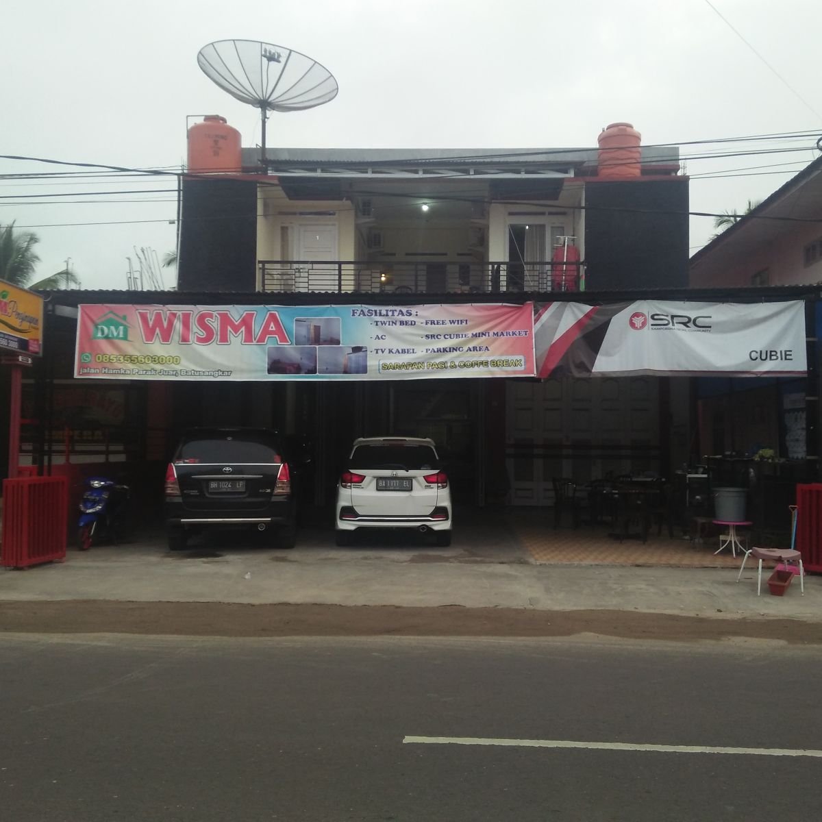 A two-story building is shown, featuring a spacious outdoor area with car parking in front. A large banner displays facility information and a satellite dish is mounted on the roof. The entrance is framed by red railings, adding a defined outline to the space.