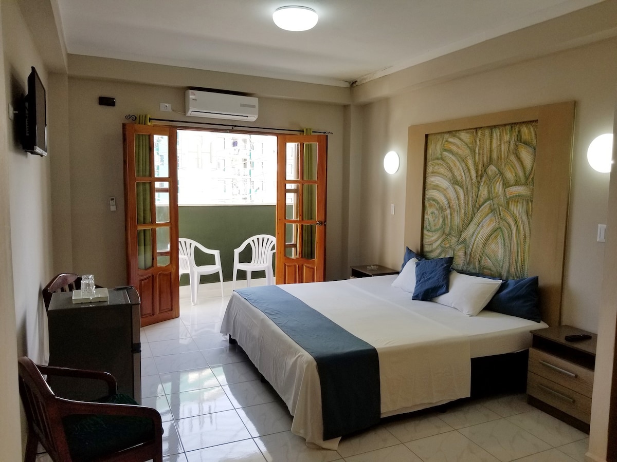 A spacious bedroom features a large bed with white bedding and decorative pillows. Two sliding glass doors lead to a private balcony with two white chairs. Soft lighting from wall fixtures enhances the room's neutral tones. A small bedside table and a wooden chair are also present.