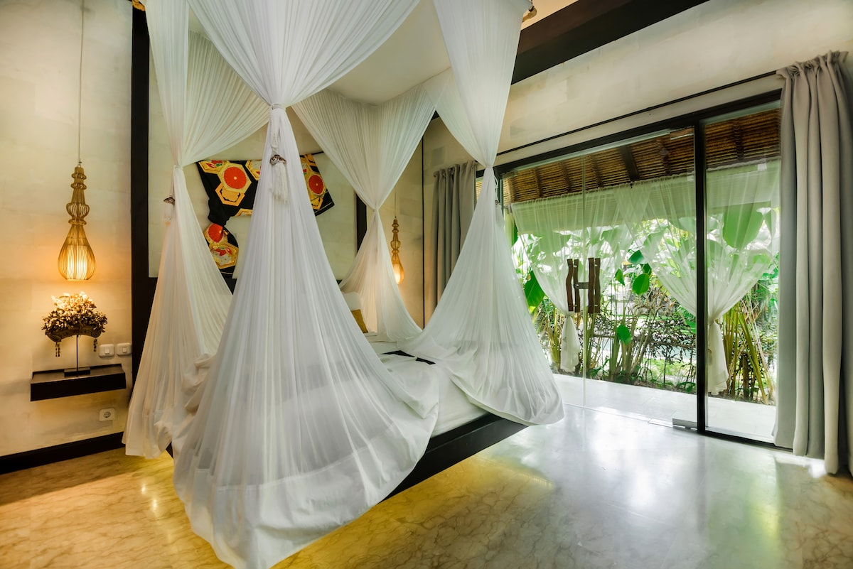 A spacious bedroom features a king-sized bed draped with flowing white curtains. Natural light enters through floor-to-ceiling glass doors, leading to a lush outdoor view. Decorative elements include a vibrant artwork and elegant pendant lighting, providing a serene and inviting environment.
