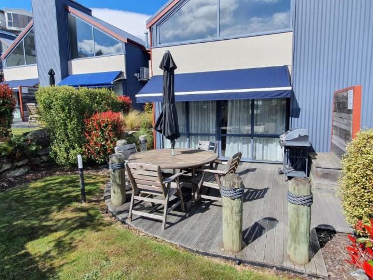 An outdoor deck area is featured, equipped with a round wooden table surrounded by matching chairs. A black umbrella provides shade, while a barbecue grill is positioned nearby. Lush greenery and colorful shrubs add natural beauty to the space.
