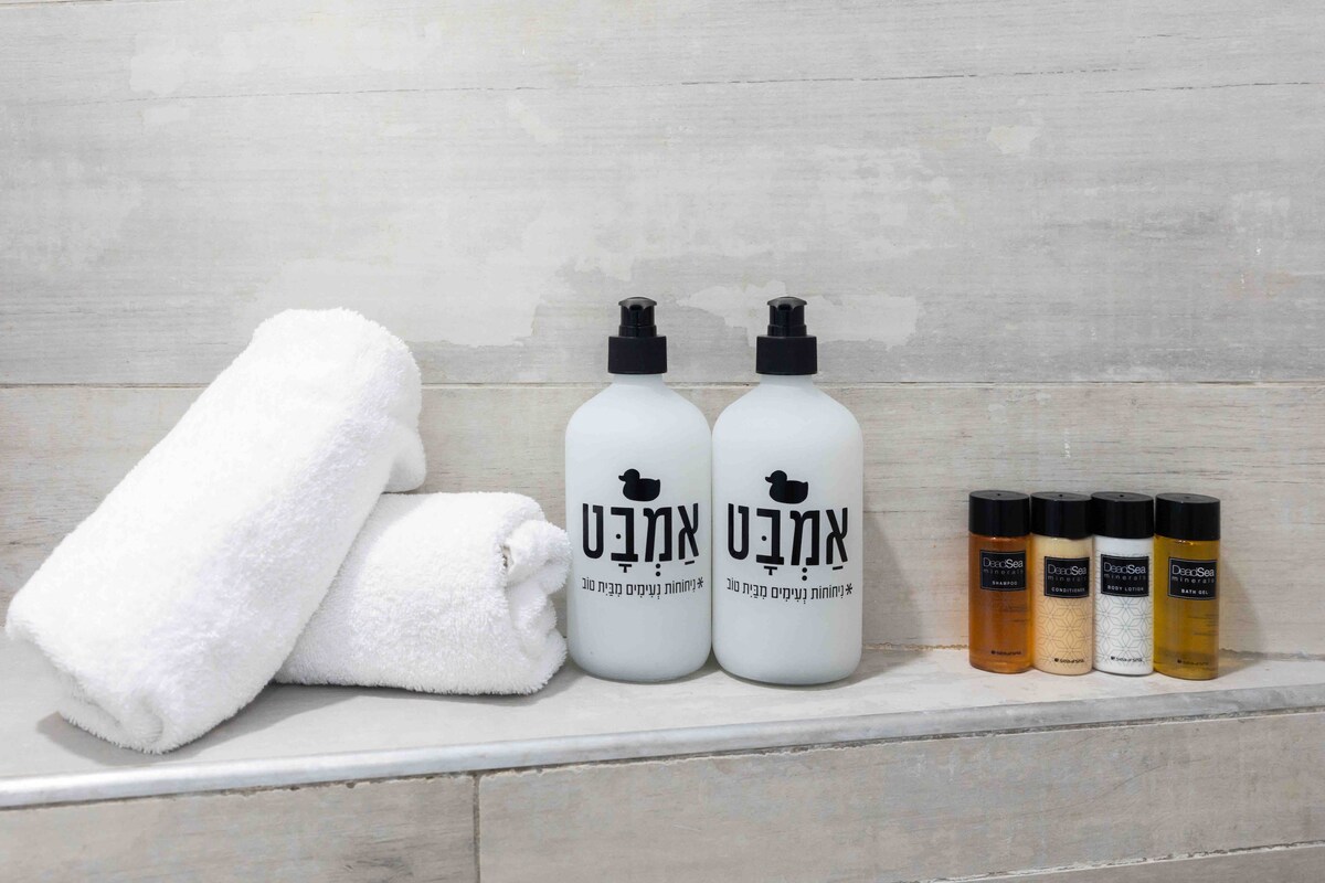A clean and minimalistic bathroom setup features two large bottles of liquid soap and shampoo in white containers with printed labels. Soft white towels are neatly arranged beside them, along with individual travel-sized toiletries in various shades of brown and clear.
