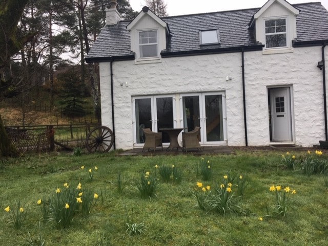 The exterior of a charming two-story cottage features a white washed facade and a slate roof. Two sets of French doors open to the patio area, which is surrounded by a garden with blooming yellow daffodils. A rustic wheel is visible nearby, enhancing the natural setting.