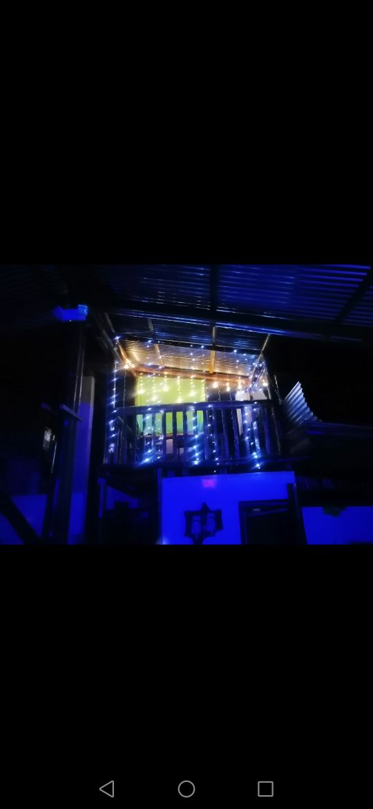 A balcony stands illuminated with warm string lights, creating a soft glow against the dark surroundings. The balcony is framed by a rustic wooden railing and appears inviting for evening relaxation.