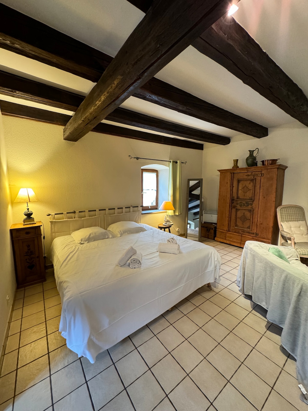 A bright bedroom features a spacious bed with white linens, complemented by a wooden dresser and a classic armchair. Exposed wooden beams enhance the room's character. A bedside lamp provides soft lighting, while a window allows natural light to illuminate the space.