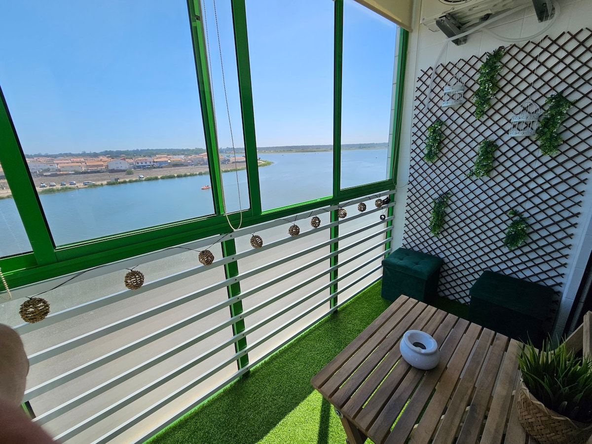 A bright balcony features large windows offering a picturesque view of the lake. The area includes a wooden table and two green stools, adorned with greenery on the walls. Decorative lights provide a soft ambiance, enhancing the inviting outdoor space.