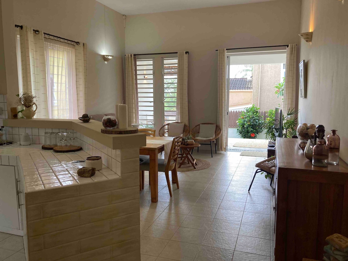 The open-concept living area features a dining table with wooden chairs and a small kitchen counter. Light filters through large windows, illuminating the space. An inviting sitting area with woven chairs is positioned for relaxation, while a small garden is visible through the sliding door.