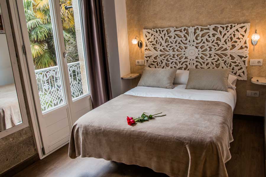 A spacious bedroom features a decorative headboard, two side tables with reading lights, and a neatly made bed. Light enters through glass doors that open to a small balcony, revealing greenery outside. A single rose rests on the bedspread, adding a touch of color.