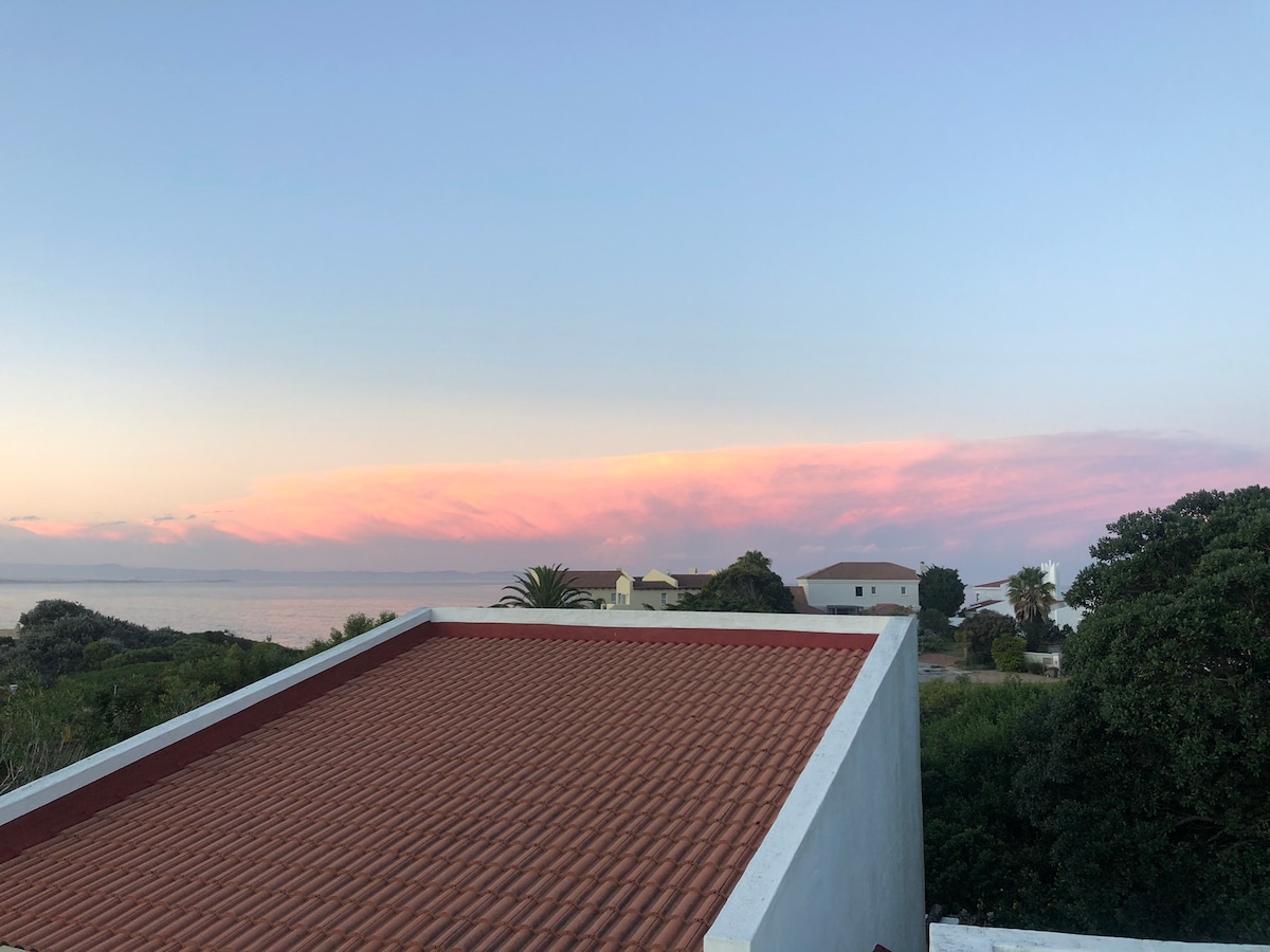 A serene view captures a sunset sky adorned with soft pink and purple hues over a tranquil ocean. The image features rooftops and greenery in the foreground, contributing to a peaceful coastal landscape.