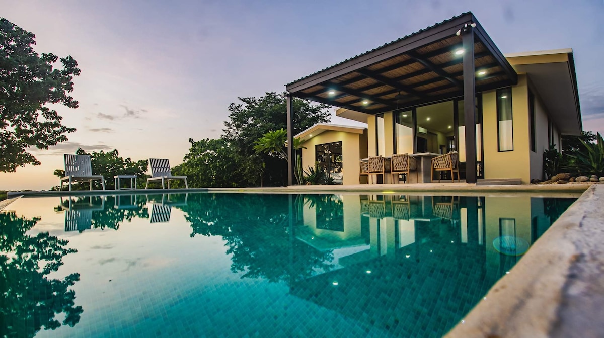 A serene outdoor space features a reflective swimming pool bordered by lounge chairs and tropical greenery. The modern house, with large outdoor overhangs, complements the tranquil atmosphere, while the sky transitions to evening hues, enhancing the natural beauty of the landscape.