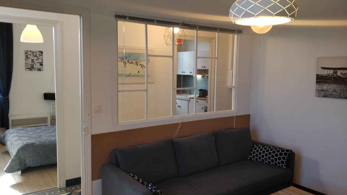 A spacious living area is highlighted by a cozy sofa with patterned cushions. Natural light filters through a window that opens to the kitchen area. The room features contemporary light fixtures and a neutral color palette, creating a welcoming environment.