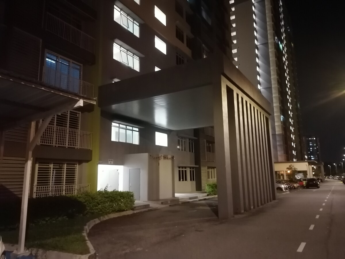 The apartment entrance is shaded by a modern canopy supported by vertical pillars. Soft lighting illuminates the building's facade, highlighting the contrasting colors of the walls. Parking spaces are visible, providing easy access for guests arriving by car.