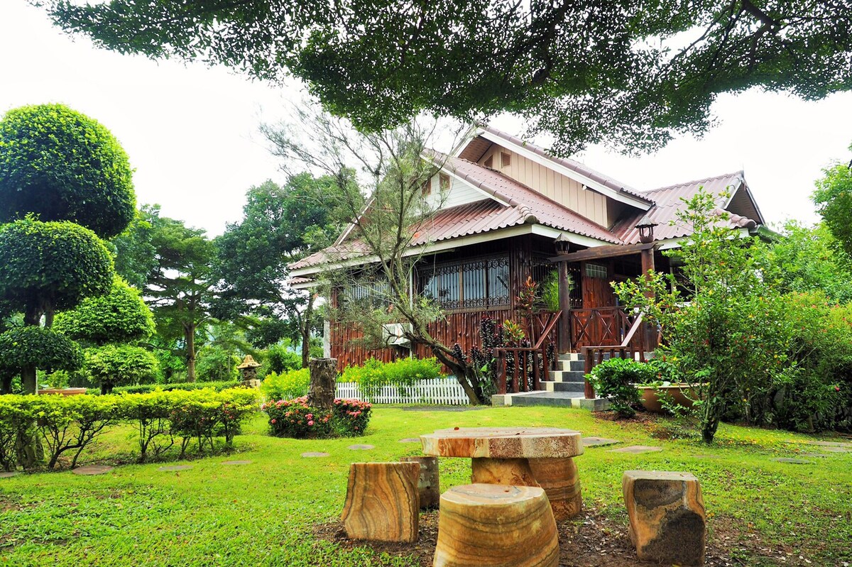 A charming wooden house is set among lush greenery, featuring a wraparound porch and large windows. The surrounding garden is meticulously landscaped, with neatly trimmed shrubs and stone seating arranged on the lawn, inviting relaxation in a serene outdoor setting.