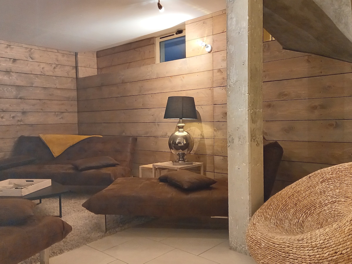 The living area features wooden paneling and soft, earthy tones. Lounge seating is arranged around a low table. A lamp stands on a side table, casting a warm glow. Textured cushions and a woven chair add to the inviting environment.